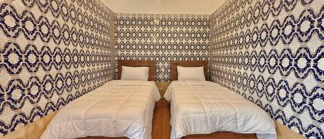A room with two beds, wooden flooring, patterned walls, and a ceiling light.