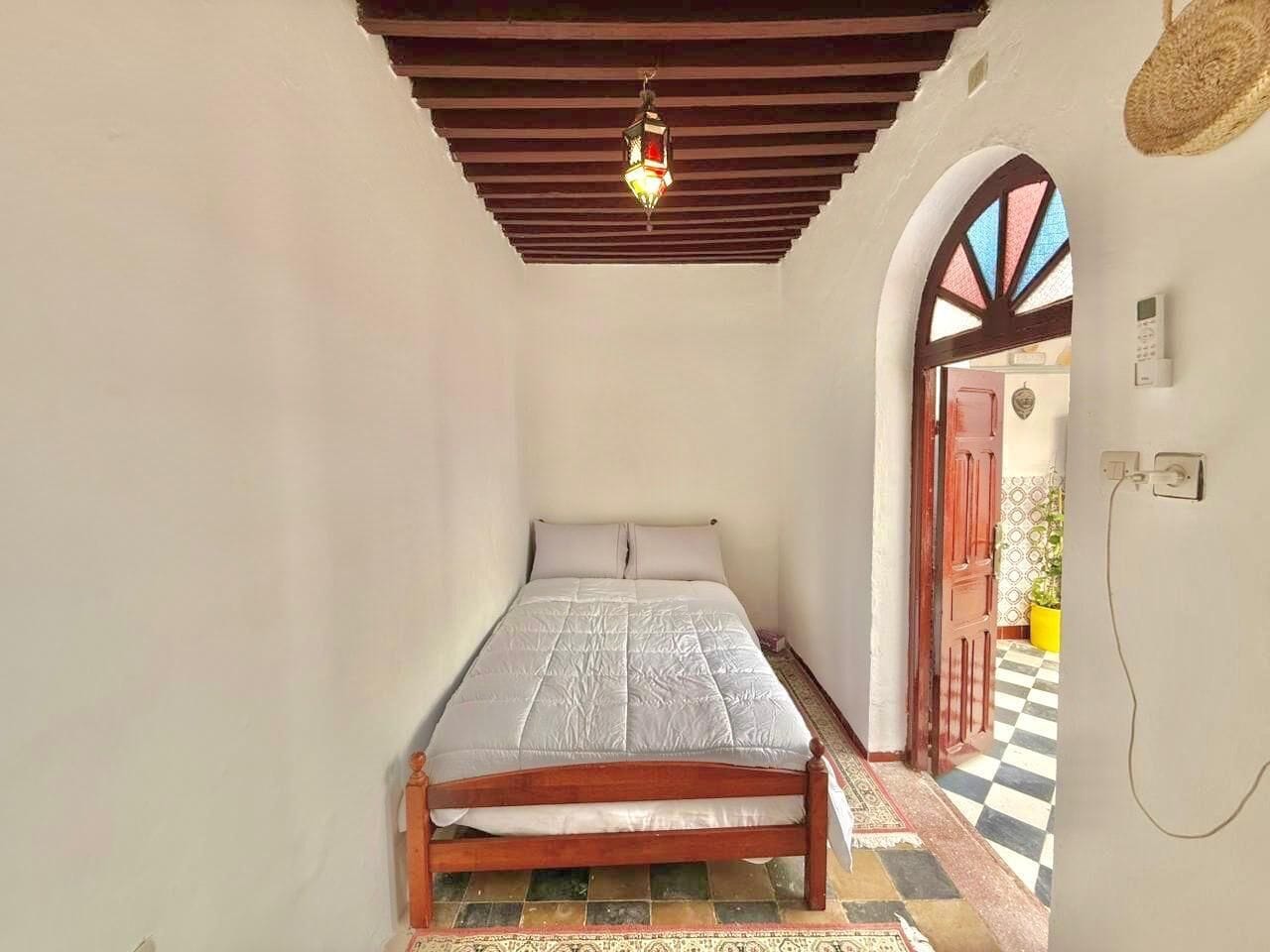 A small, single-bed room with a wooden bed frame, a hanging light fixture, a checkered floor, and a doorway leading to an outdoor area.