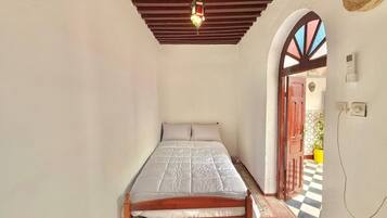 A small, single-bed room with a wooden bed frame, a hanging light fixture, a checkered floor, and a doorway leading to an outdoor area.