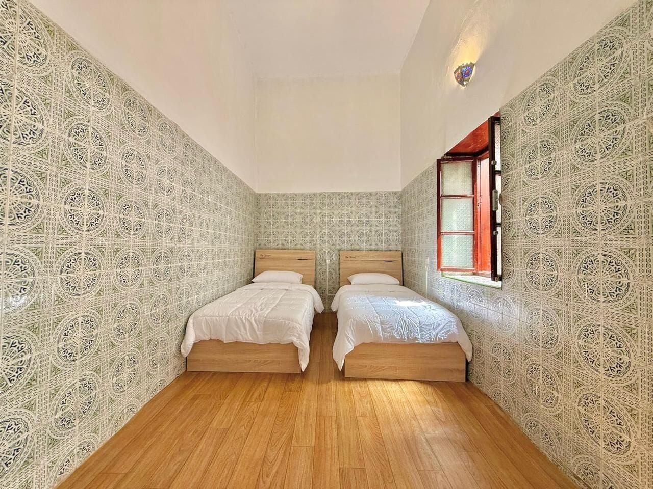 Habitación con dos camas, suelo de madera, paredes con diseño y una ventana con marco rojo.