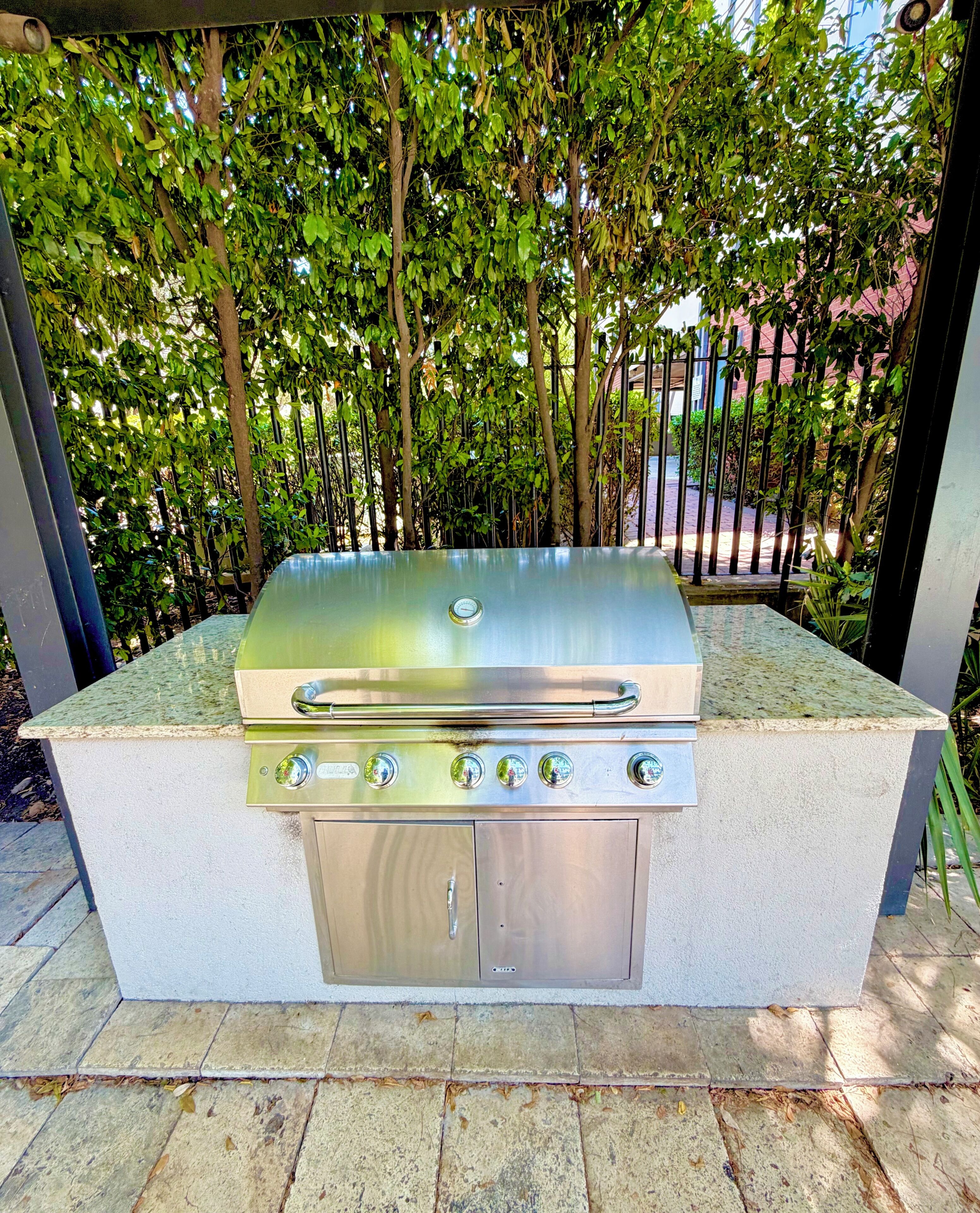 Una barbacoa d'exterior amb acabat d'acer inoxidable, quatre cremalls i un panell de control, amb fons de verd vegetació i una tanca metàl·lica.