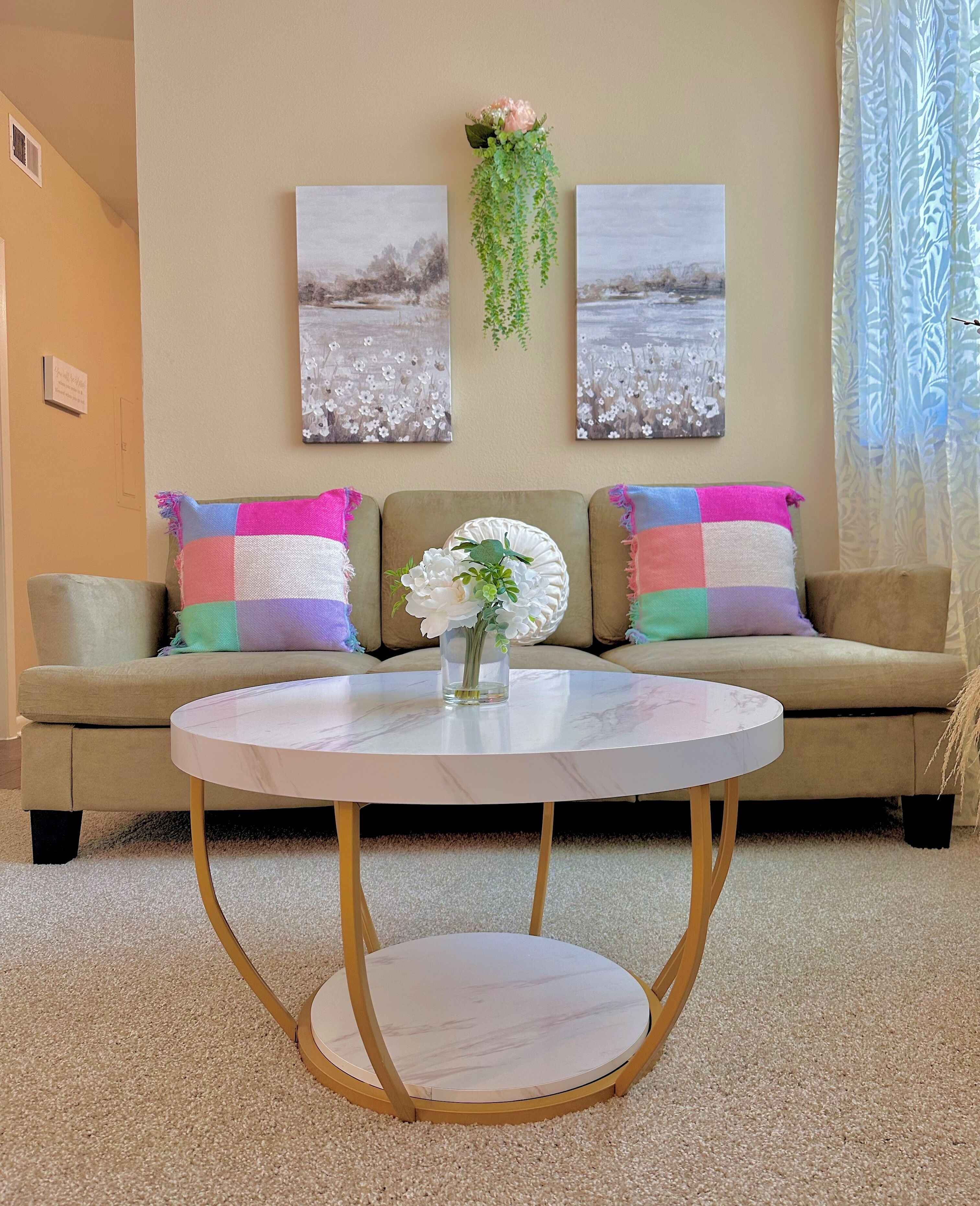 A living room with a beige sofa, colorful pillows, a round marble coffee table, and two floral wall art pieces above the sofa.