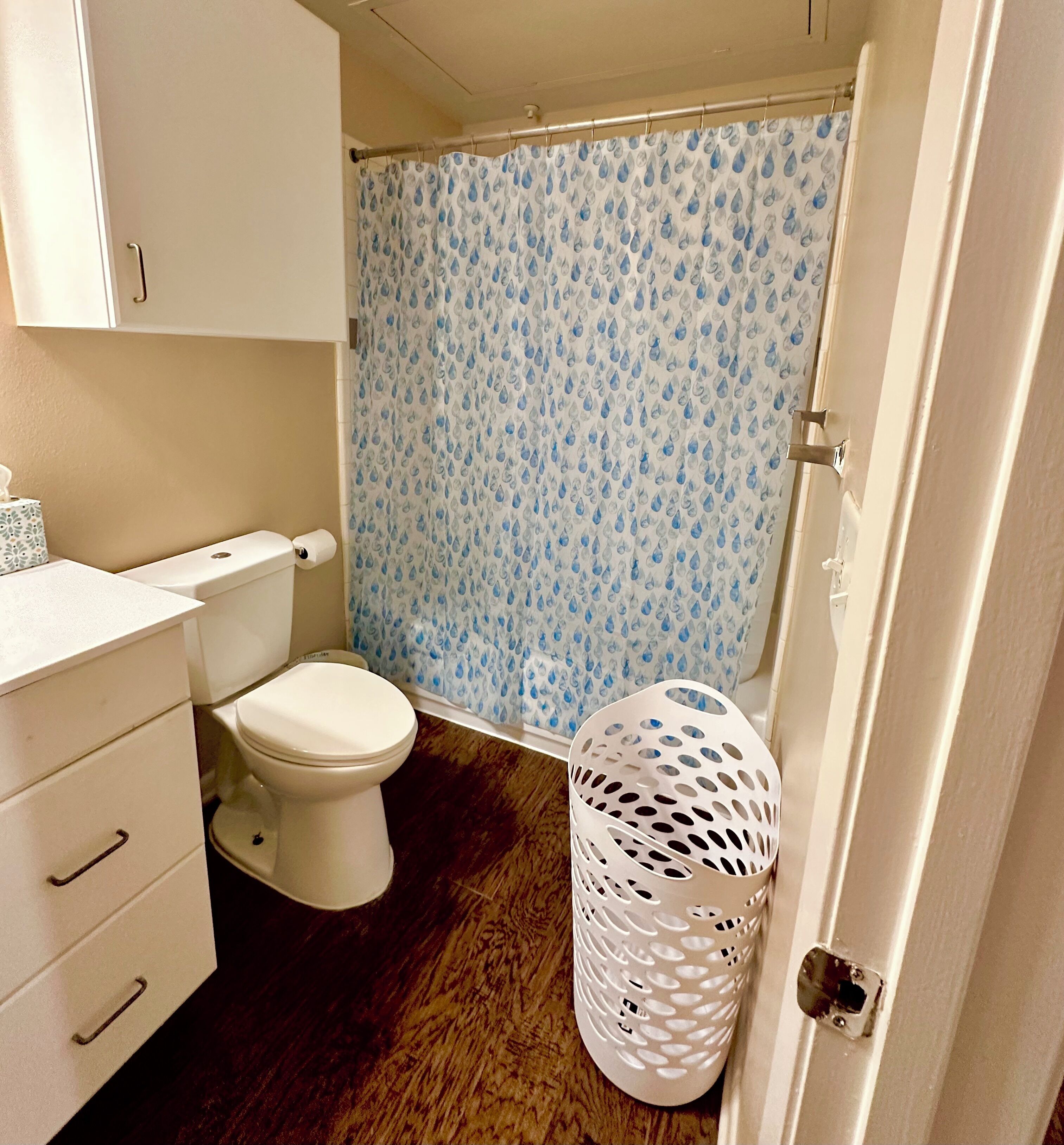 A bathroom with a toilet, a shower curtain with a water droplet pattern, a white laundry basket, and a white cabinet with drawers.