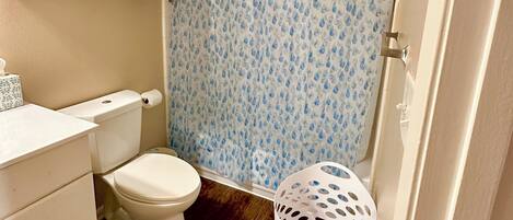 A bathroom with a toilet, a shower curtain with a water droplet pattern, a white laundry basket, and a white cabinet with drawers.