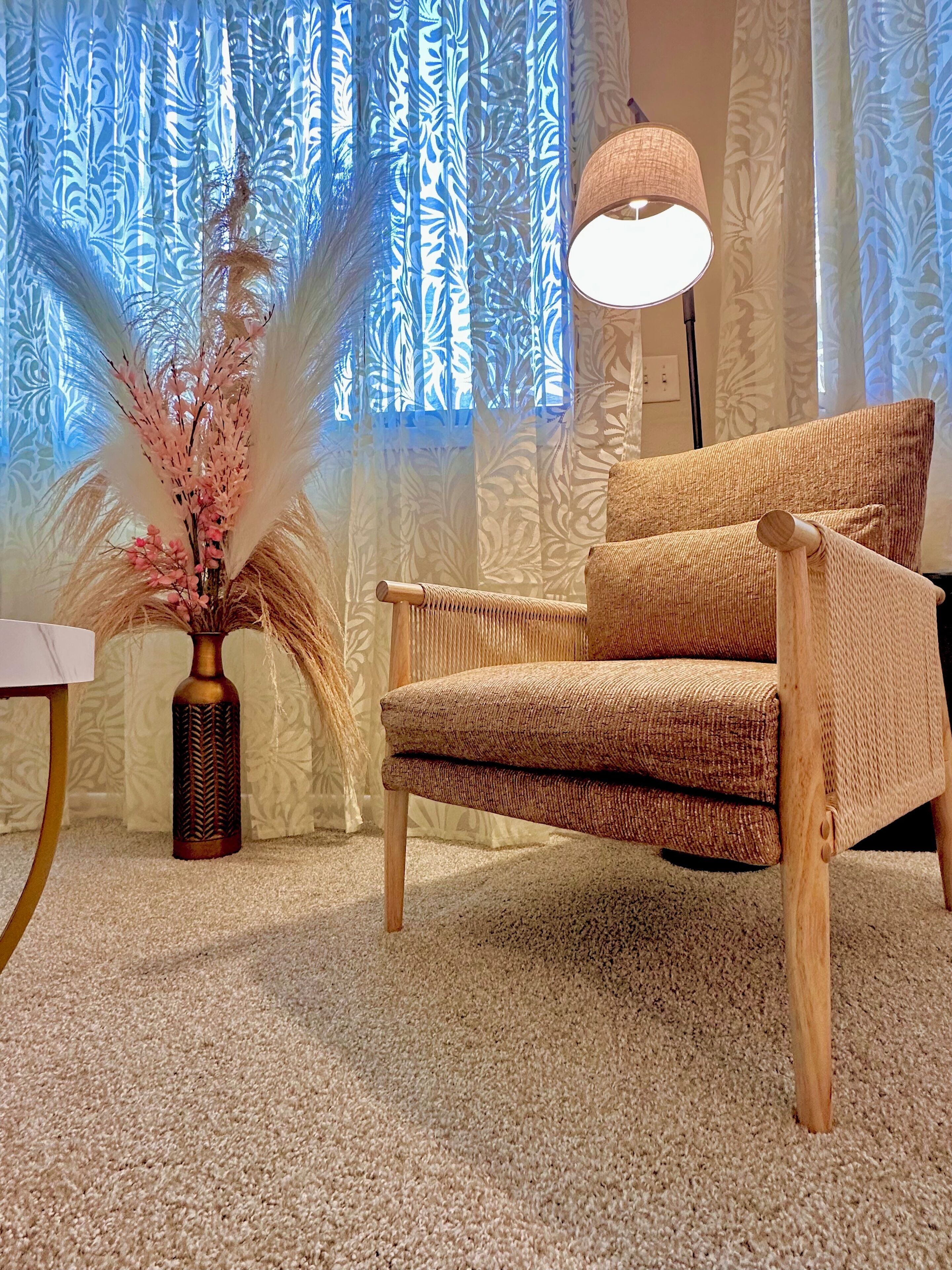 A cozy room with a woven armchair, a vase with dried flowers, and a lamp.