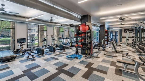 Ein großzügiger Fitnessraum mit unterschiedlichen Trainingsgeräten, großen Fenstern und einem Schachbrettmuster auf dem Boden.