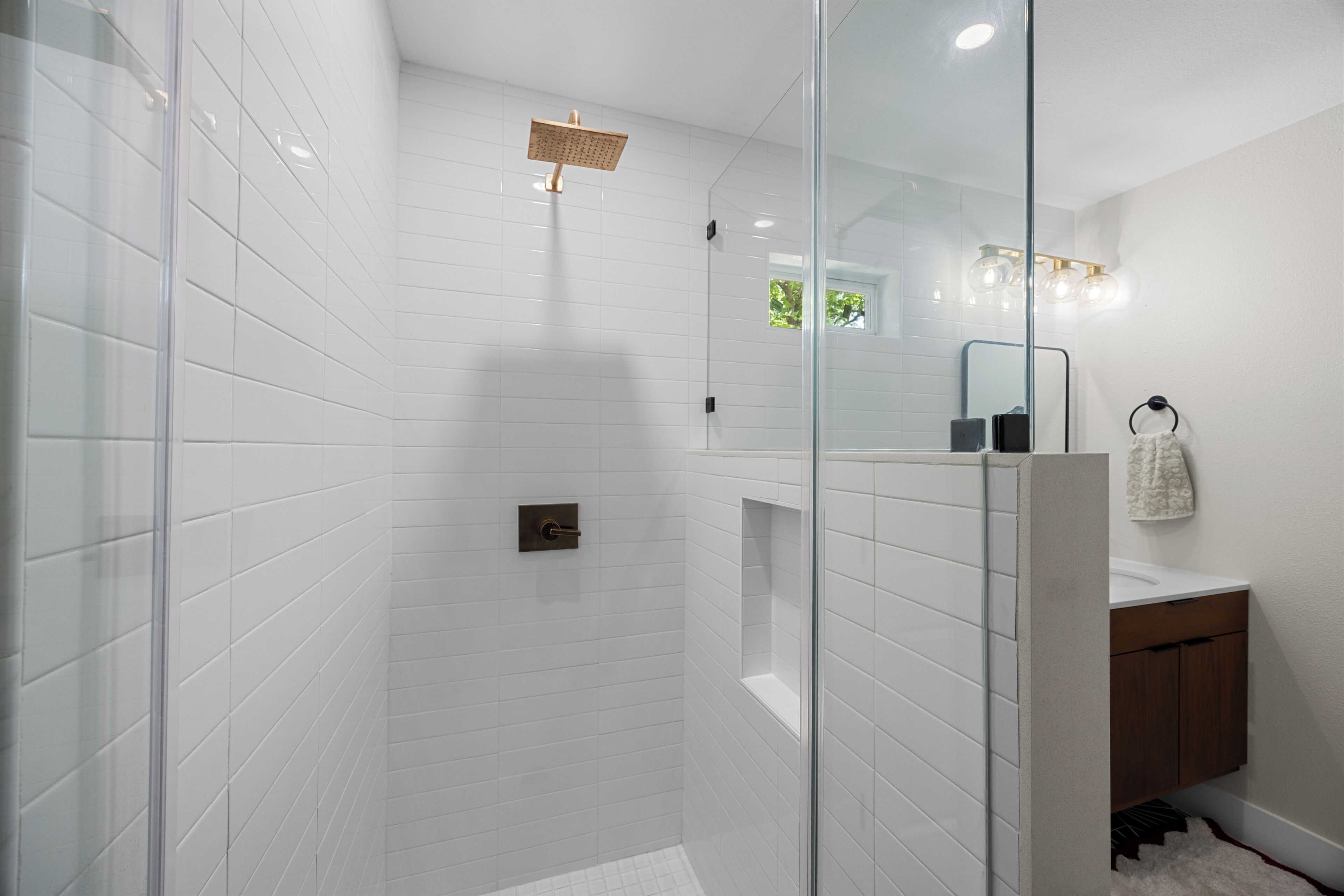 Un baño moderno con azulejos blancos, cabina de ducha de vidrio, tocador de madera y espejo con iluminación.