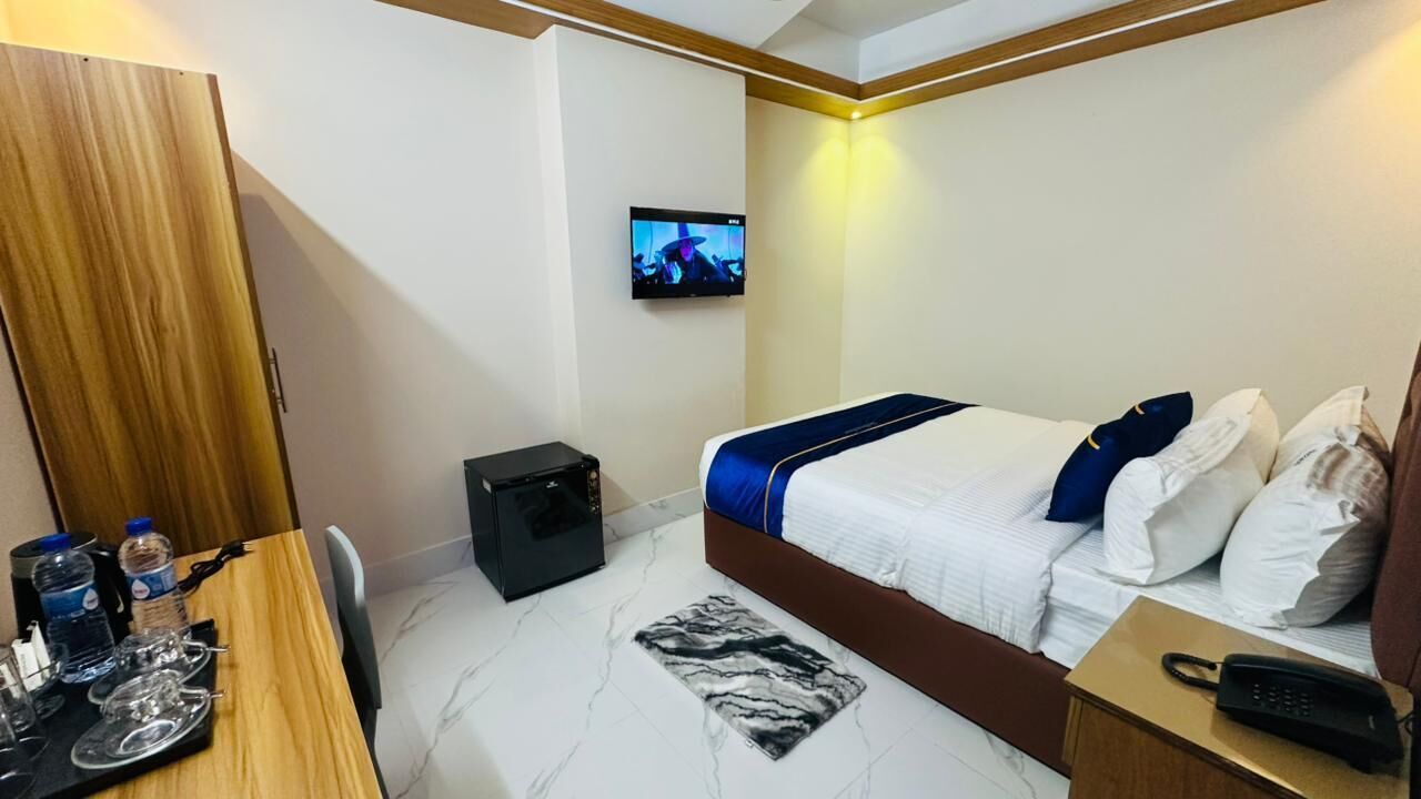 Une chambre d’hôtel avec un lit, un téléviseur fixé au mur, un bureau avec une chaise et une petite table sur laquelle se trouvent une bouteille d’eau et des verres.