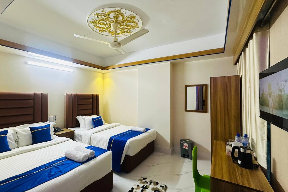 A hotel room with two beds, a ceiling fan, a flat-screen TV, a small trash bin, and a wooden side table with a kettle and cups.