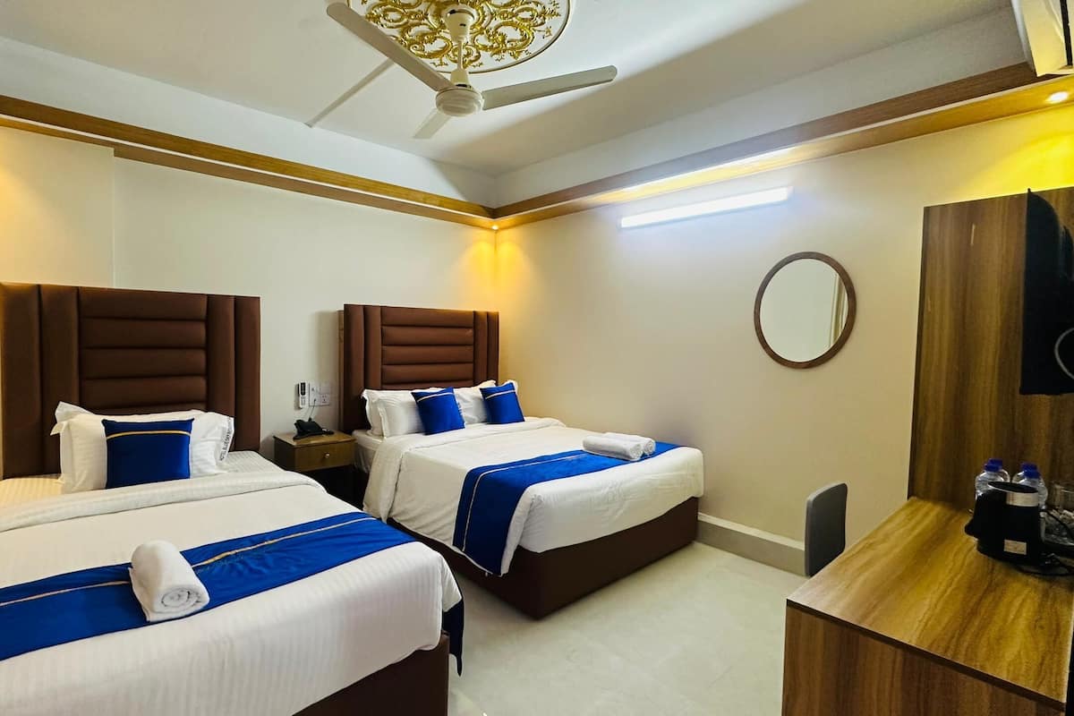A hotel room with two beds, each with a brown headboard, white bedding, and blue accents. There is a wooden desk with a mirror and a bottle of water.