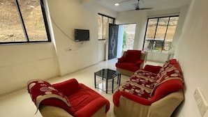 A living room with two sofas, a glass coffee table, and a red armchair. There is a TV mounted on the wall and a ceiling fan.