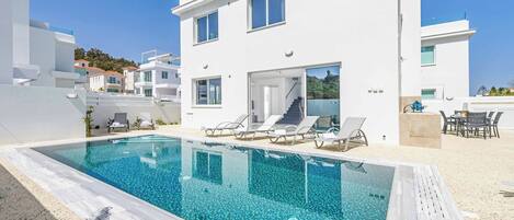A modern white villa with a swimming pool, sun loungers, and a view of the surrounding area.