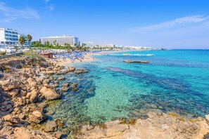 Une plage avec une eau turquoise et limpide, un rivage rocheux et plusieurs bâtiments visibles au loin.