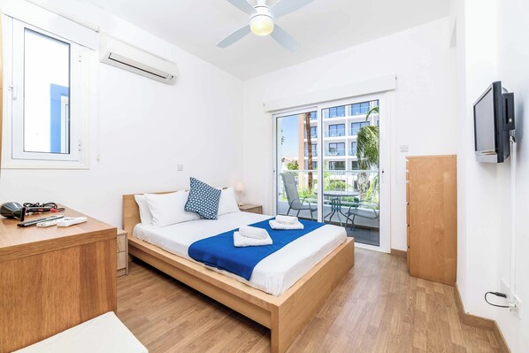 A modern hotel room with a bed, a desk, a TV, and a balcony with a view of buildings and trees.