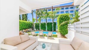 A modern outdoor lounge area with wicker furniture, a glass coffee table, and a view of a swimming pool and building in the background. - Villa Atlantis - Three Bedroom Villa, Sleeps 6 (Protaras)