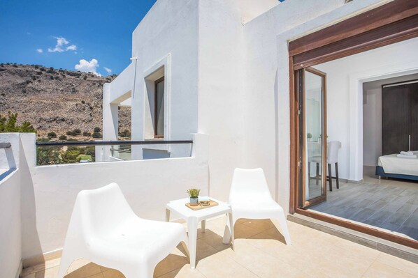 A balcony with white plastic chairs and a small table, a bedroom with a bed and a sliding glass door, and a mountainous landscape in the background.