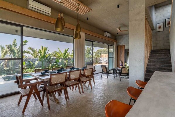 A spacious dining area with a large table and chairs, large windows with a view of palm trees, and a staircase leading to an upper level. - Villa Maaya (Chikkaballapur)