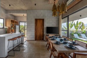 A modern kitchen with a long dining table set for six, a bar area with stools, a TV, and a large window with a view of the outdoors. - Villa Maaya (Chikkaballapur)