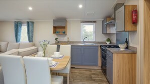 A modern kitchen with grey cabinets, a wooden countertop, and a built-in oven. There is a dining area with a table and chairs, and a window with blue curtains. - Marton Mere Holiday Village - 17890 (Blackpool)