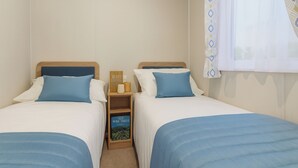 A twin room with two beds, each with a blue headboard and pillow, a bedside table with a book, and a window with a patterned curtain. - Marton Mere Holiday Village - 17890 (Blackpool)