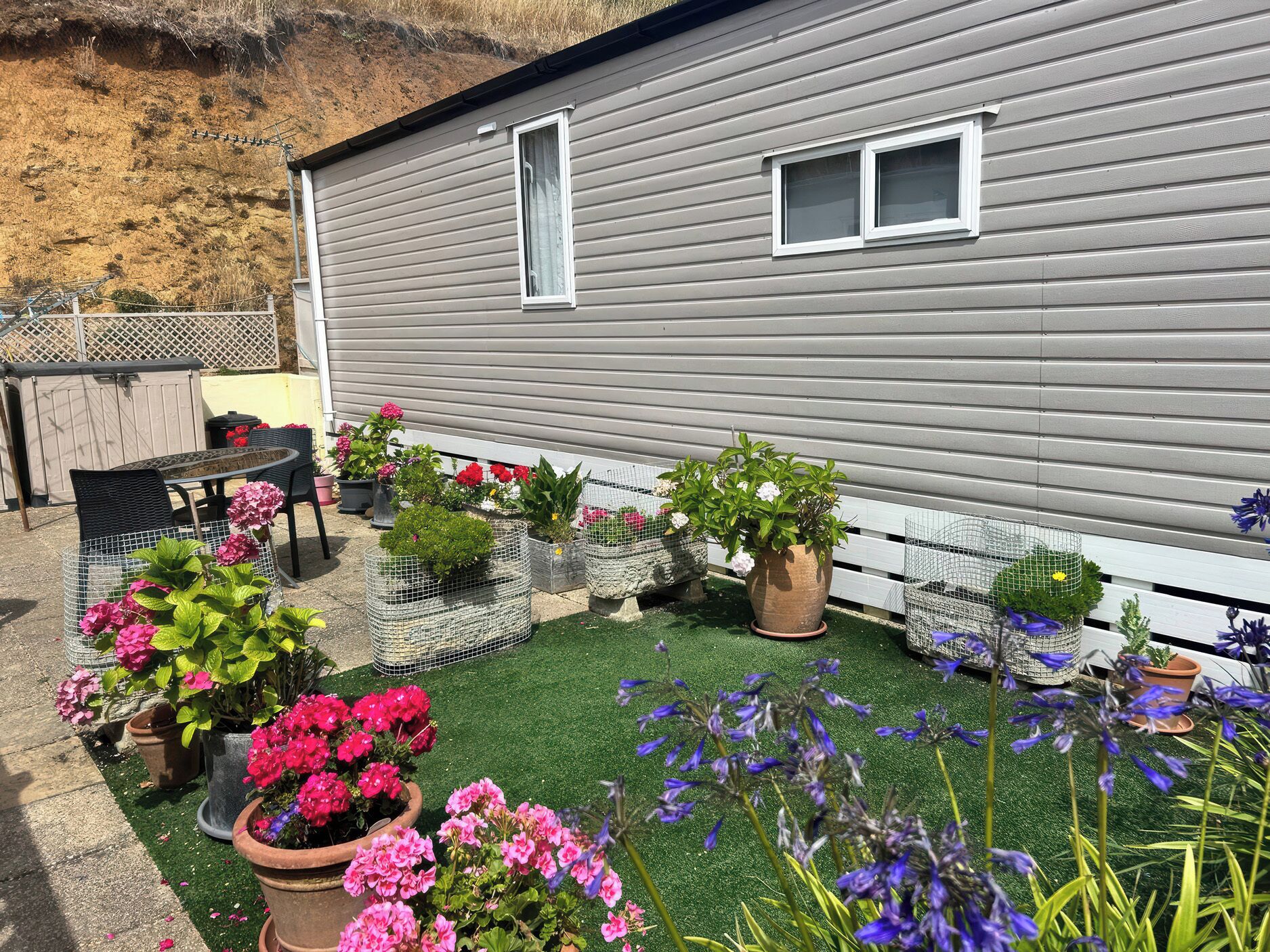 Una propiedad residencial con una casa gris, un patio con mesa y sillas, y varias macetas con plantas y flores.
