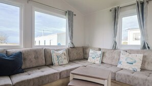 Un salon chaleureux avec un canapé d’angle beige, une table basse en bois et des coussins à motifs. De grandes fenêtres offrent une vue sur l’extérieur.