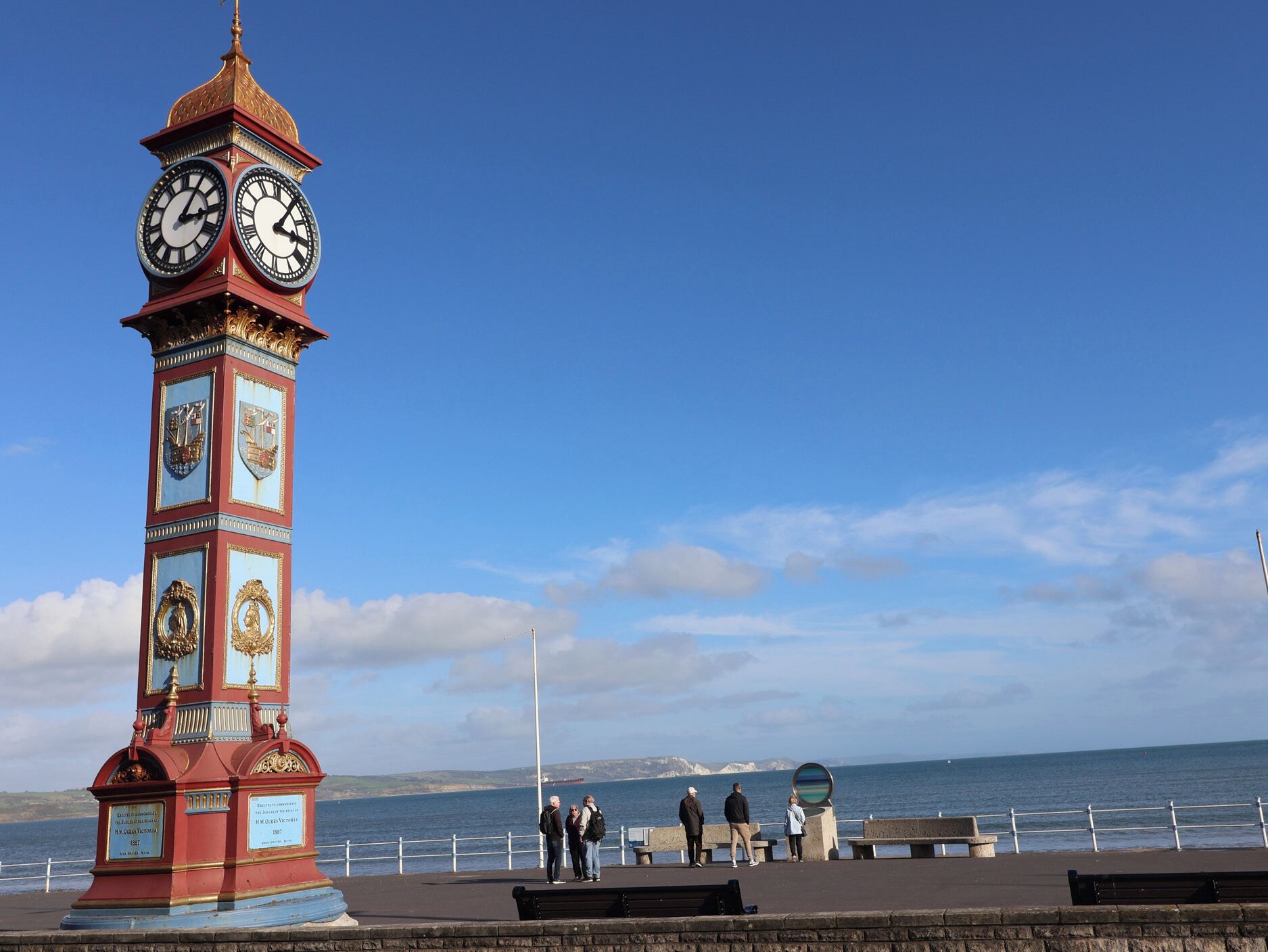 A clock tower with a plaque, a coastal view, and a promenade with benches.