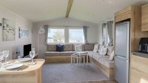 Une pièce chaleureuse et bien éclairée, avec une table à manger en bois mise pour deux personnes, un canapé confortable avec des coussins, une petite table blanche et un réfrigérateur.