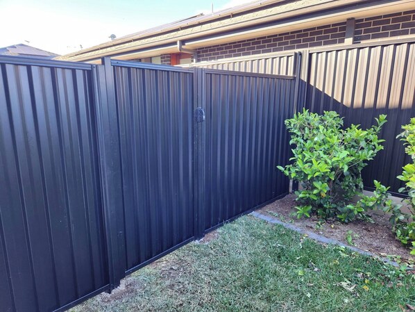 A black metal fence with a gate, a small bush, and a brick building in the background. - Couples' Haven - 2 Bedrooms (Sydney)
