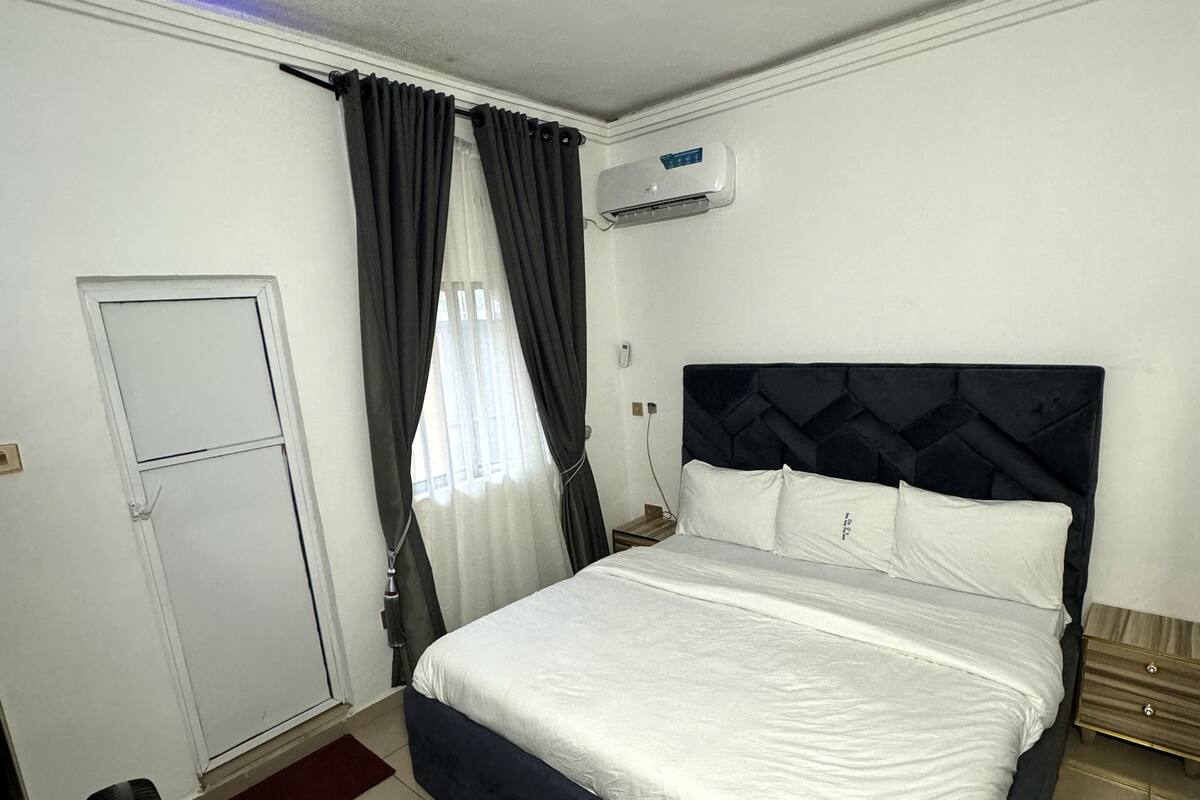 A bedroom with a large bed, a ceiling fan, an air conditioning unit, and a door.