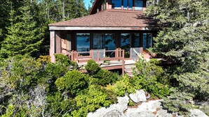 A two-story wooden cabin with large windows, surrounded by dense forest.