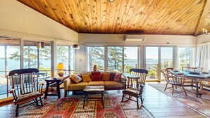 A spacious living area with a wooden ceiling, large windows offering a view of the outdoors, a sofa, chairs, and a dining table set for a meal.