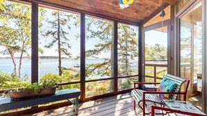 A covered porch with a bench and a table, overlooking a body of water and trees.