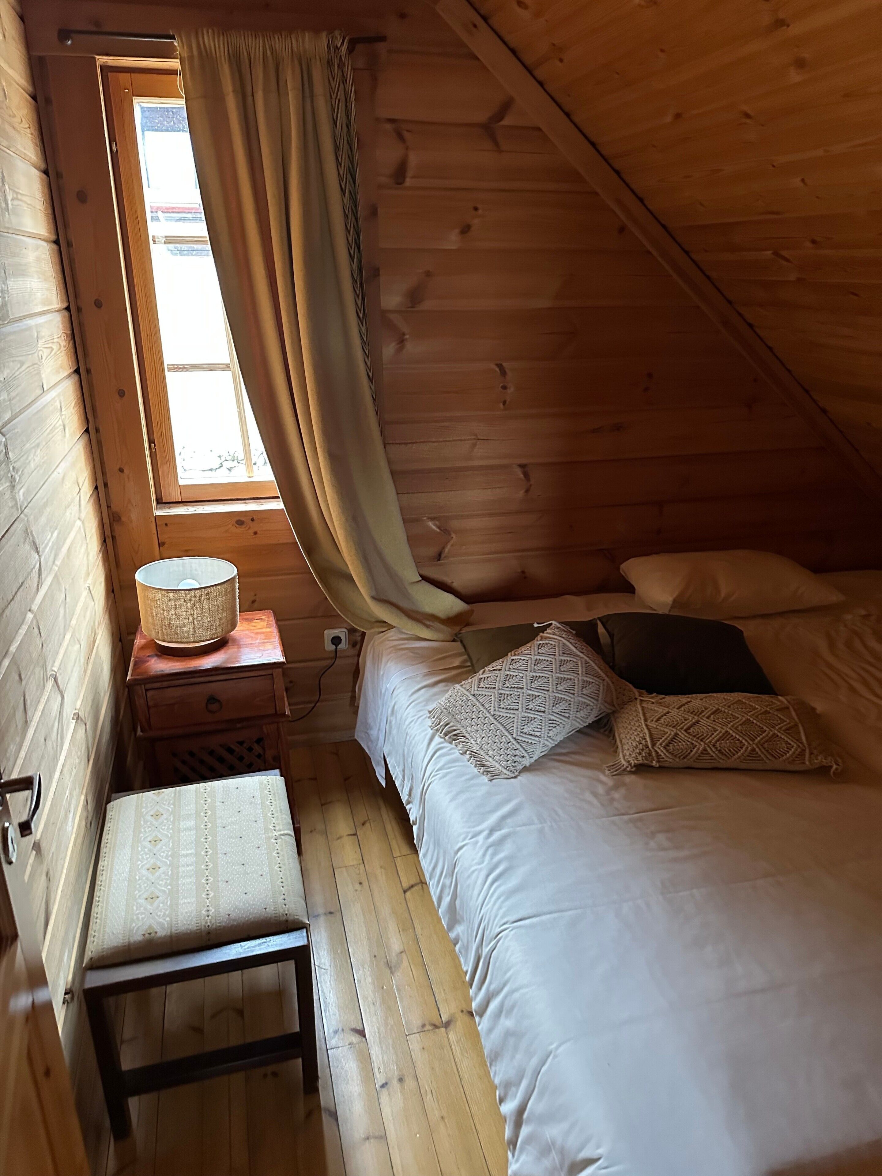 Et hyggeligt soveværelse i skunken med skråvægge, en enkeltseng med mønstrede puder, et natbord med lampe og et vindue med gardiner.