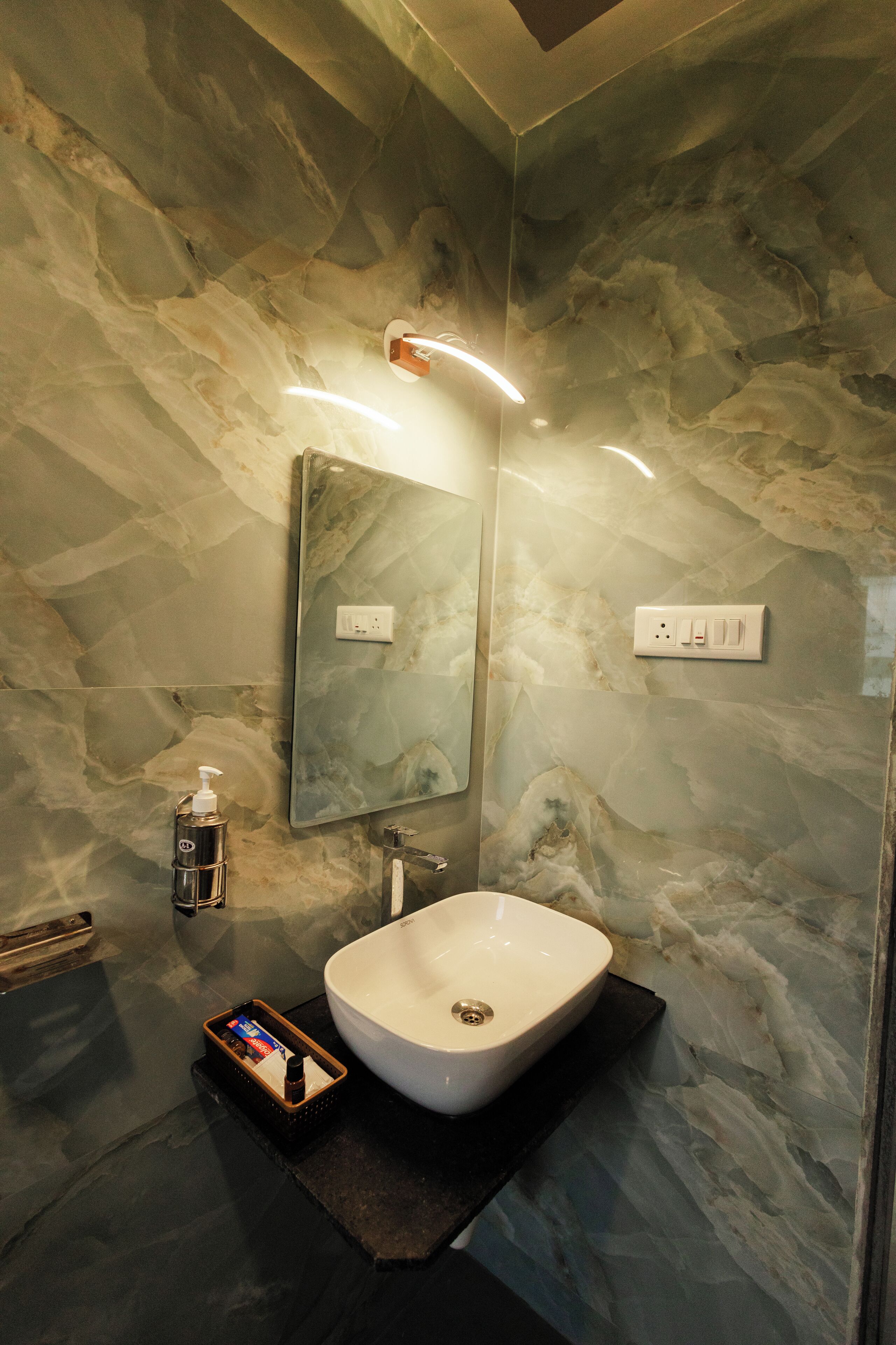 Een moderne badkamer met een marmeren wand, een grote spiegel, een witte wasbak en een zeepdispenser.