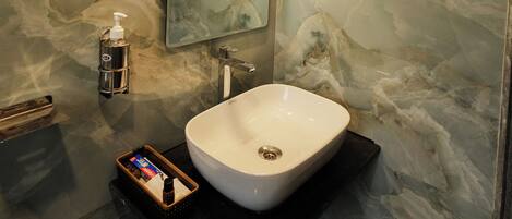A modern bathroom with a marble wall, a large mirror, a white sink, and a handheld soap dispenser.