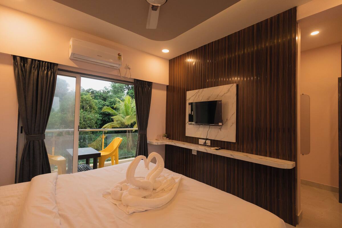 A hotel room with a bed, a TV mounted on the wall, a balcony with a view of greenery, and an air conditioning unit.