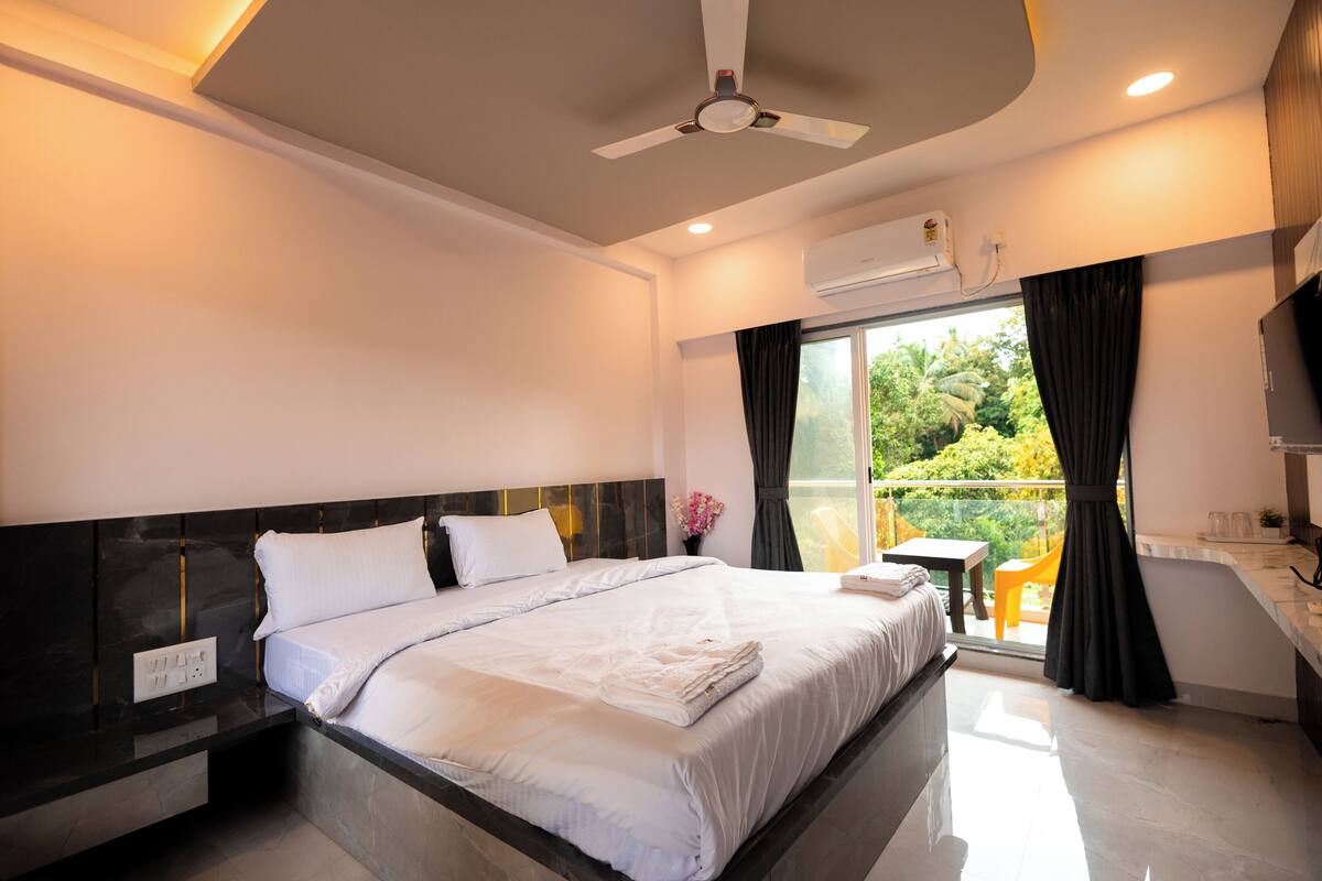 A modern hotel room with a large bed, a ceiling fan, an air conditioning unit, a small balcony with a table and chairs, and a TV mounted on the wall.