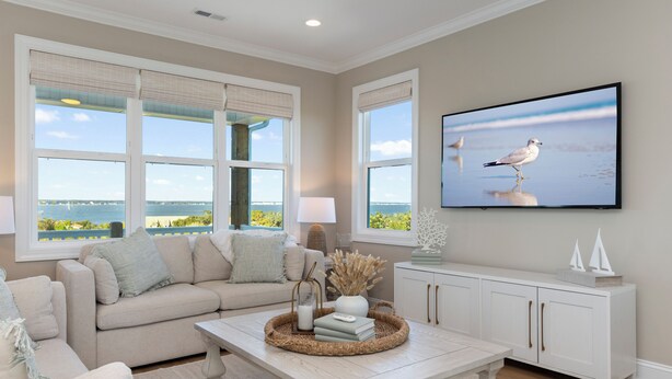 A living room with a beige sofa, a coffee table with decorative items, a television mounted on the wall, and large windows with a view of the sea. - Endless Views | Luxury 4BR Emerald Isle Retreat w/ Pool & Ocean Breezes (Emerald Isle)