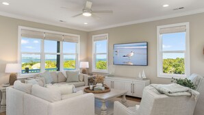A spacious living room with a large window, a ceiling fan, and a flat-screen TV. - Endless Views | Luxury 4BR Emerald Isle Retreat w/ Pool & Ocean Breezes (Emerald Isle)