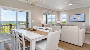 A spacious living room with a white dining table, white chairs, a beige sofa, and a large window offering a view of the sea. - Endless Views | Luxury 4BR Emerald Isle Retreat w/ Pool & Ocean Breezes (Emerald Isle)