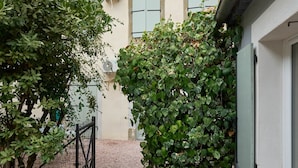 A courtyard with a gravel path, a metal gate, a building with light-colored walls and shutters, and a tree with green leaves.