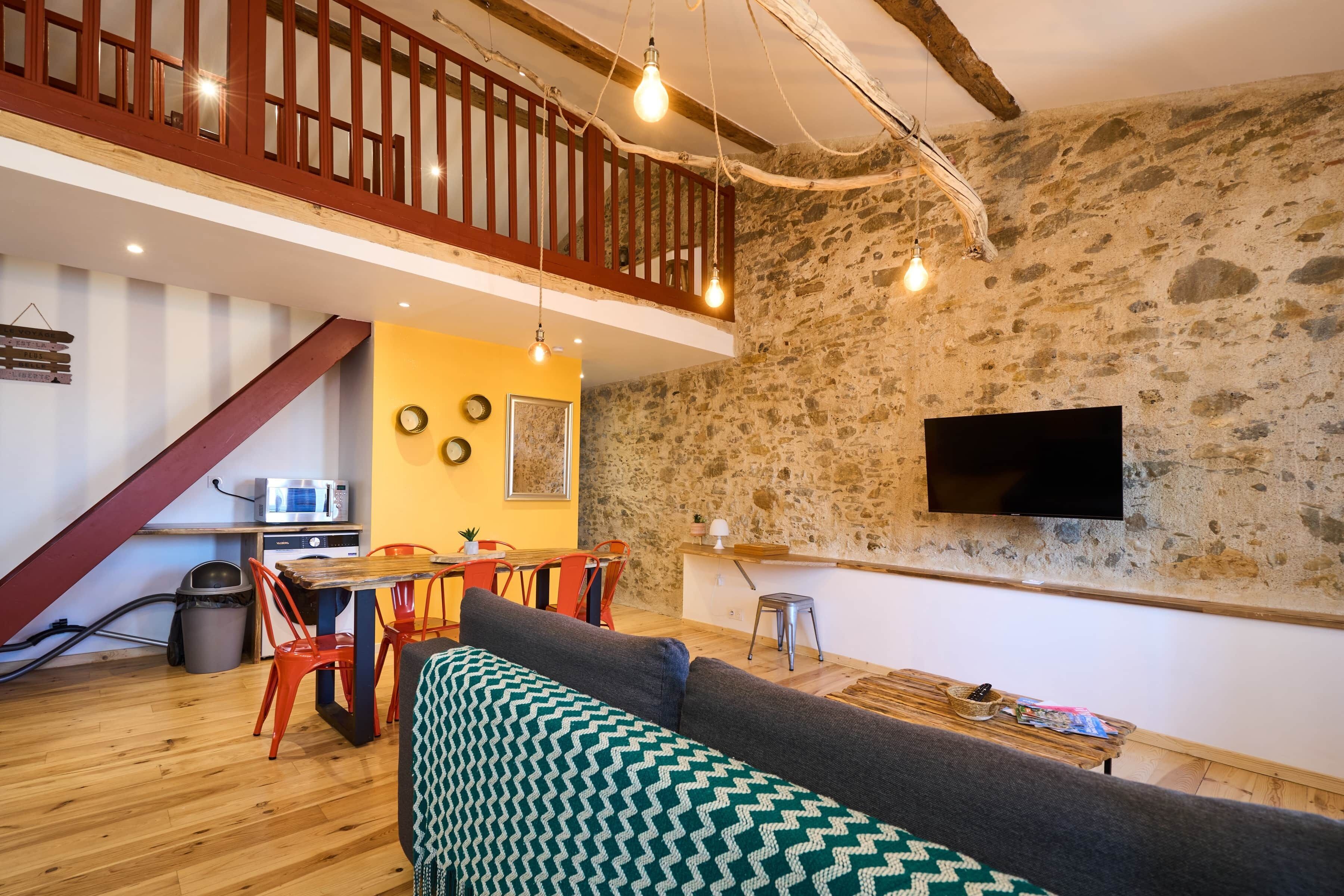 A cozy living area with a wooden floor, a sofa, a dining table with chairs, a TV mounted on a stone wall, and a staircase with a wooden railing.