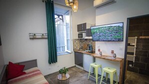 Un espacio habitable compacto con una cama, una pequeña zona de comedor con taburetes, una cocina con televisor y una ventana con cortinas.