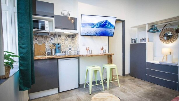 Cocina compacta con salpicadero de azulejos, un refrigerador pequeño, un televisor montado en la pared y dos taburetes.