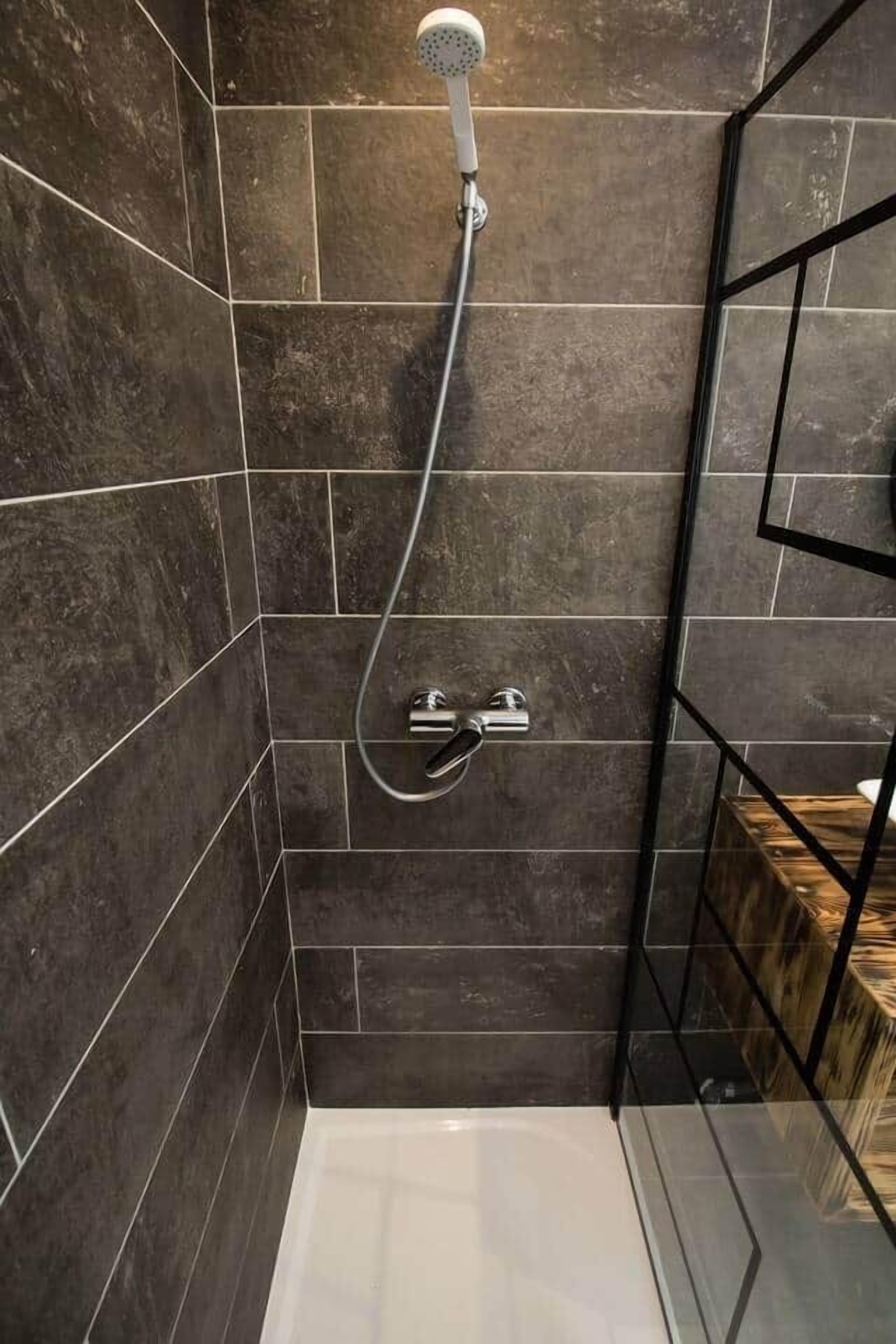 A modern shower with dark tiles, a glass enclosure, and a wooden storage unit.