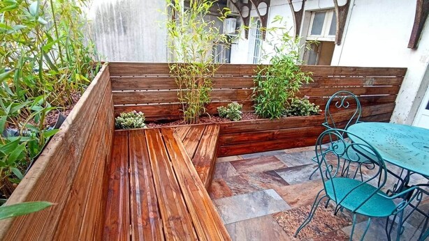 Un banco de madera y una mesa de metal con sillas en un jardín delimitado por una valla de madera y con diversas plantas.