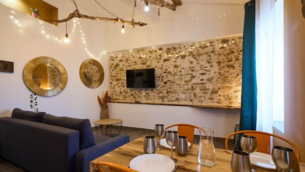 A dining area with a rustic stone wall, a mounted TV, and string lights. - Apartment 4 pers Calme & Confort - Clim - Centre (Carcassonne)