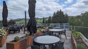 Un patio al aire libre con mesas y sillas redondas, cubierto por sombrillas, con vistas a un paisaje pintoresco.