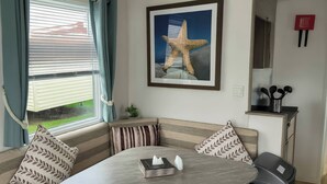 Een kamer met een ronde tafel, gestreepte kussens, een kunstwerk van een gestileerde zeester, een raam met zonwering en een klein kastje met keukengerei.
